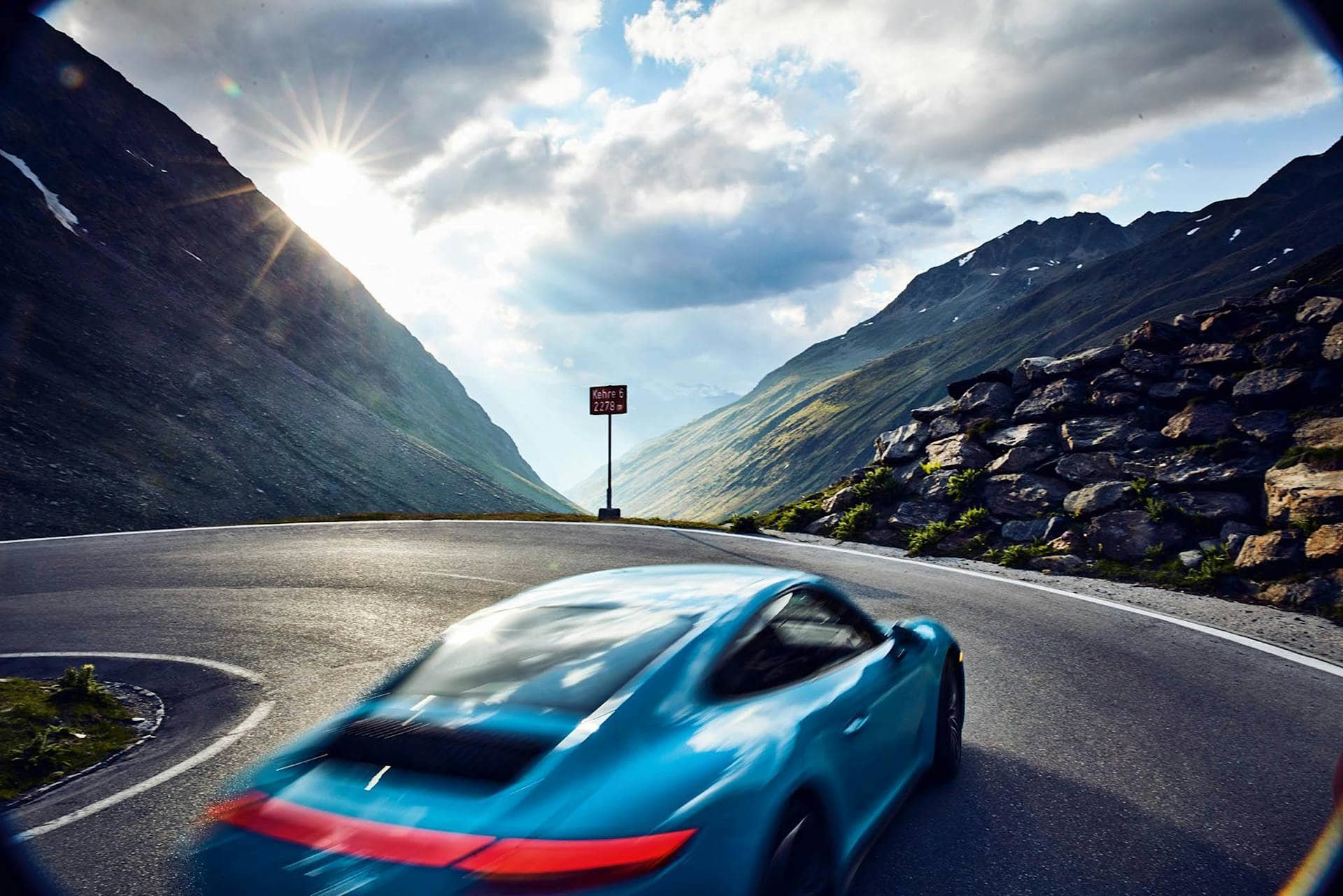 Blue Porsche Speeds Around Mountain Curve With Sunburst Background, St